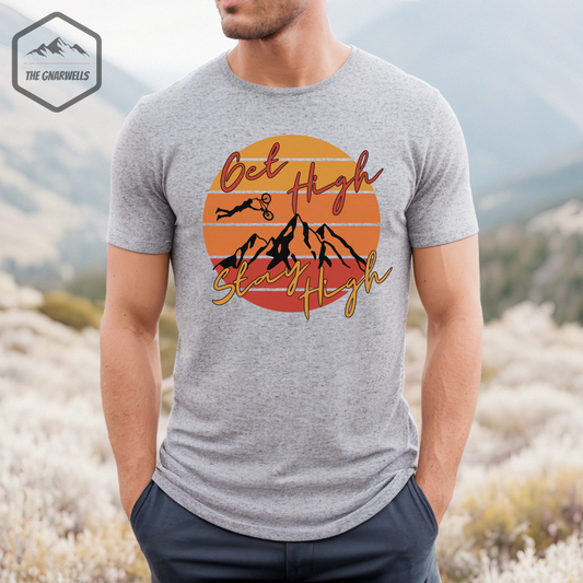 The Get High Stay High mountain bike shirt with a circle shape subset into the background with sunset colors. There is a mountain range in the foreground with the words get high stay high.